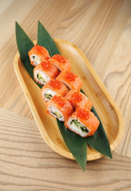 Suşi ruloları bambu tabakta güzelce düzenlenir. Her rulo somon, salatalık ve turuncu balık yumurtası ile kaplı. Ayar, doğal ışığı olan ahşap bir masa..
