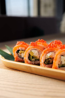 Sebze ve balıklarla dolu suşi ruloları ahşap bir tabakta dikkatlice düzenlenir. Turuncu havyar tepeyi süslüyor, restorandaki görsel cazibeyi arttırıyor..