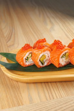 Somon ve avokadoyla dolu suşi ruloları üzerinde balık yumurtası olan ahşap bir tabağa yerleştirilmiş. Taze malzemeler gündelik yemek ortamında Japon mutfağını ön plana çıkarıyor..