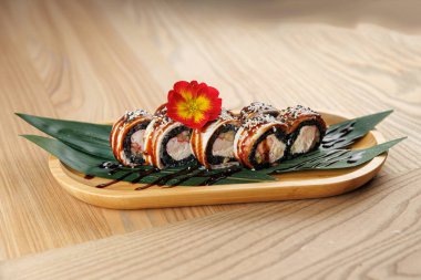 Suşi yuvarlakları ahşap bir tepside, kırmızı bir çiçekle süslenmiş ve yeşil yapraklara yerleştirilmiş. Suşide çeşitli malzemeler yer alıyor. Restoran ortamında Asya mutfağı sergileniyor..