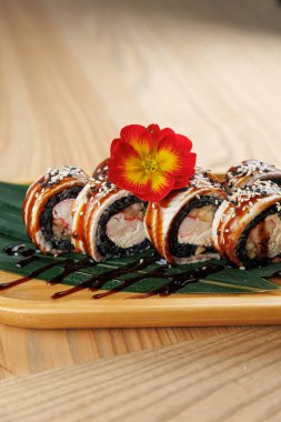 Suşi ruloları ahşap bir tabakta, pirinç, deniz yosunu ve deniz ürünleri gibi malzemeler içeriyor. Renkli bir çiçek kenarda oturur ve sunumu güçlendirir..