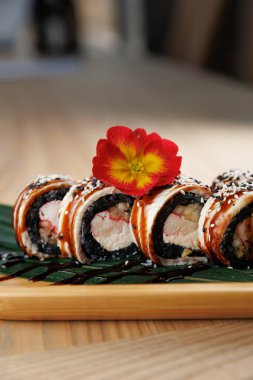 Deniz ürünleriyle dolu suşi ruloları bambu tabakta düzgünce düzenlenir. Canlı bir çiçek sunuma dekoratif bir dokunuş ekler. Dekor, çağdaş bir yemek havası veriyor..