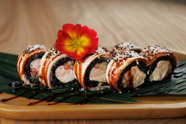 Taze suşi ruloları, susam tohumları ve serpiştirilmiş sosla ahşap bir tabakta güzelce düzenlenir. Renkli bir çiçek sunuma renk katar.