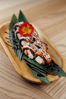 Suşi ruloları yeşil yapraklarla kaplanmış, çiçeklerle süslenmiş ve sosla süslenmiş ahşap bir tabakta düzenlenir. Sahne rahat bir restoranda geçiyor..