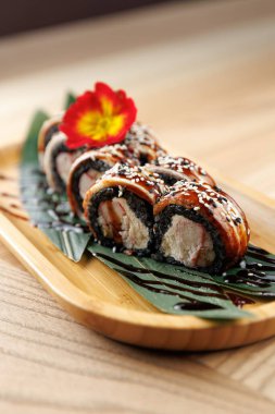 Suşi ruloları bambu tabakta güzelce düzenlenir. Tabakta siyah pirinç, deniz ürünleri ve dekorasyon için bir çiçek garnitürü yer alıyor. Sıradan bir yemek ortamında servis edilmiş..