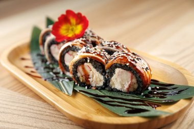 Suşi rulolarında yengeç, krem peynir ve susam tohumları ve çiçekle süslenmiş siyah pirinç var. Tabak ahşap bir tabakta yeşil yapraklarla sunuluyor. Paylaşmak için ideal..