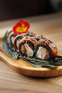 Suşi ruloları ahşap bir tabağa düzgünce yerleştirilir. Tabakta çeşitli malzemeler yer alıyor ve bir çiçekle süsleniyor. Dekorlar rahat bir restoran atmosferi sergiliyor..