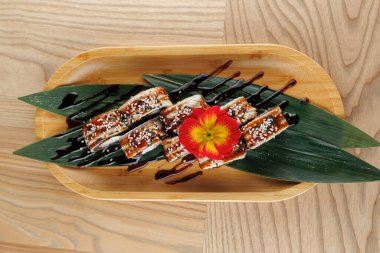 Sushi dürümleri, susam tohumları ve çiçeklerle süslenmiş bambu bir tabakta güzelce düzenlenir. Tabak yeşil yapraklar ve yemek alanında bir sos yağmuru ile tamamlanır..