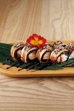 Çeşitli dolgularla suşi ruloları ahşap bir tabakta zarif bir şekilde oturur. Bir çiçek biraz renk katar. Ortam modern ve gündelik, çeşitli mutfaklardan zevk almak için mükemmel..