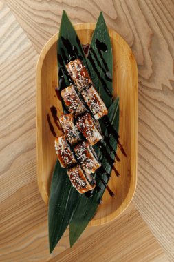 Suşi ruloları taze bambu yaprakları üzerine yerleştirilir, susam tohumları ve zengin bir sosla süslenir. Ayarda sunumu geliştiren ahşap bir tablo yer alıyor.