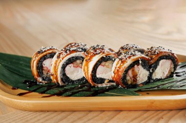 Sushi rolls are arranged neatly on a wooden plate, featuring rice, seaweed, and various fillings. Green leaves add a touch of freshness. A dark sauce is drizzled over the top.