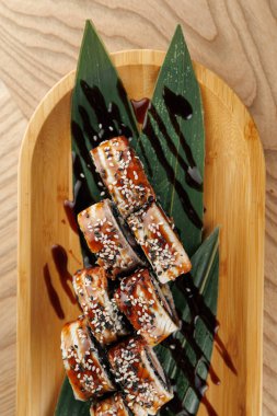 Yılanbalığıyla yapılan suşi ruloları ve üzerine susam tohumları ahşap bir tepside sunulur. Kara sos çiselemesi yeşil yaprakların yanında görüntüyü güçlendirir ve cezbedici bir yemek yaratır..