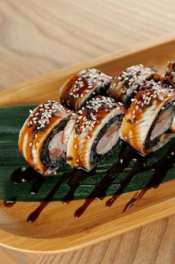 Sushi dürümleri ahşap bir tepside, susam tohumlarıyla süslenmiş ve sosla serpilmiş olarak düzenlenir. Dekor, Japon restoranlarında tipik olarak temiz ve modern bir tasarıma sahip..