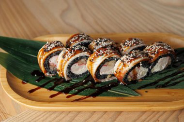 Suşi ruloları ahşap bir tabakta susam ve sos ile güzelce düzenlenir. Yeşil bir yaprak yemeği süslüyor, çekici bir sunum oluşturuyor..