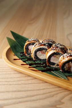 Suşi ruloları ahşap bir tabakta yeşil yapraklarla ve üzerine sos serpiştirilmiş bir şekilde düzgünce düzenlenir. Bu modern yemek sahnesinde Japon mutfağından popüler bir yemek sergileniyor..