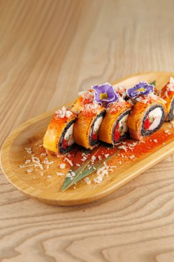 Suşi ruloları ahşap bir tabakta güzelce düzenlenir. Tabak yenilebilir çiçekler ve süslemelerle süslenmiş. Sıcak ve davetkar bir atmosfer yemek deneyimini güçlendirir..