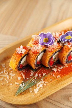 Suşi ruloları, yenilebilir çiçekler ve malzemelerle süslenmiş ahşap bir tabakta düzenlenir. Ayarlar modern bir yemek sahnesinin tipik bir mutfak sunumunu gösteriyor..