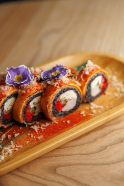 Suşi ruloları, yenilebilir çiçeklerle kaplı ve pullarla süslenmiş ahşap bir tabakta sergilenir. Sahne, Japon mutfağının bir yemek alanındaki sanatını yansıtıyor..
