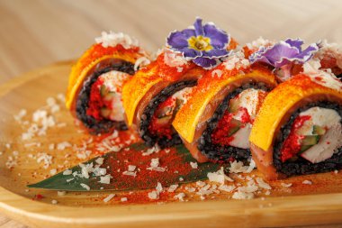 Suşi ruloları ahşap bir tabakta güzelce düzenlenir. Ekmekler sarı malzemelerle kaplı ve mor çiçeklerle süslenmiş. Tabakta baharat ve garnitür var..