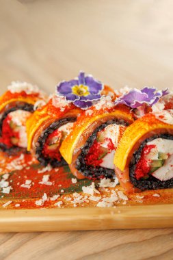 Bir grup suşi rulosu parlak renkler ve çeşitli malzemeler içeren ahşap bir tabakta düzenlenir. Yenilebilir çiçekler ve baharatlar yemeğe görsel cazibe katar..