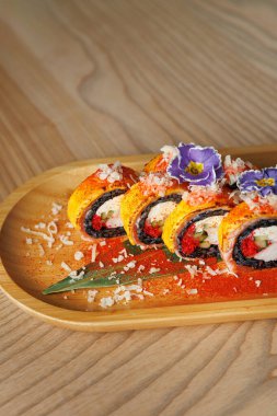 Suşi ruloları ahşap bir tabakta güzelce düzenlenir. Parlak renkler ve yenilebilir çiçekler süsleme katar. Baharat tabağın etrafına saçılmış, görsel çekiciliği arttırıyor..