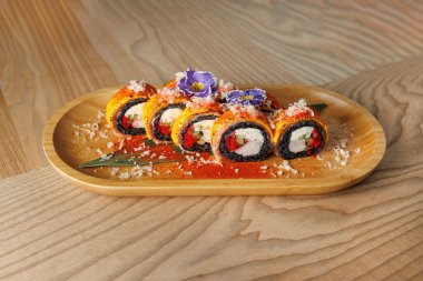 Suşi ruloları ahşap bir tabakta güzelce düzenlenir. Ekmekler canlı renklere, malzemelere ve dekoratif yapraklara sahiptir. Bu sahnede modern bir yemek atmosferi sergileniyor..