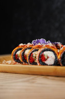 Taze malzemelerle dolu renkli suşi ruloları ahşap bir tabakta güzelce düzenlenir. Yenilebilir çiçekler sunuma sanatsal bir dokunuş ekler.