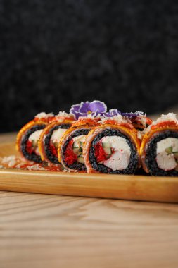 Lezzetli suşi dürümleri ahşap tabak, vitrin, canlı dolgular ve malzemeler üzerine dizilmiş. Sunum renk ve dokuların karışımını vurguluyor, bir tat deneyimini davet ediyor.