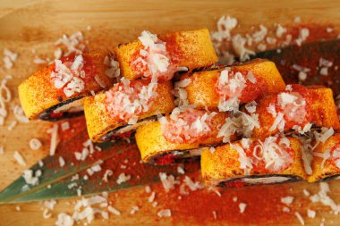 Suşi ruloları ahşap bir yüzeye istiflenmiş. Üzeri baharat ve parçalanmış malzemelerle kaplı, canlı renkler ve dokular sergileniyor. Sunuma dekoratif bir yaprak ekler.