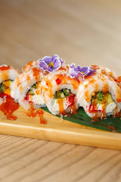 Suşi ruloları ahşap bir tabakta güzelce düzenlenir. Her rulo taze malzemelerle doludur ve parlak sos ve yenilebilir çiçeklerle kaplıdır..