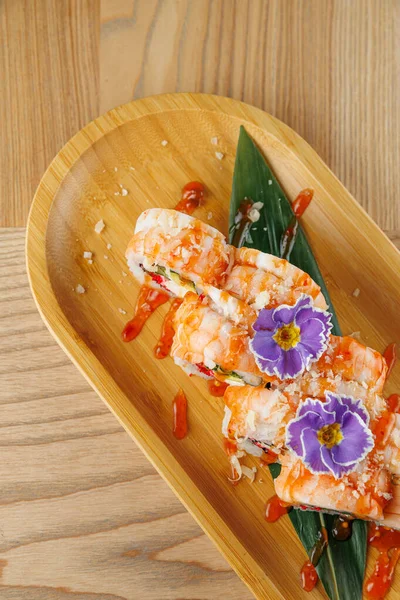 Üç suşi rulosu bir bambu tabağına konur. Her rulo karides ve renkli süslemeler içeriyor. Tabakta garnitür ve sos var. Japon mutfak sanatı sergileniyor..