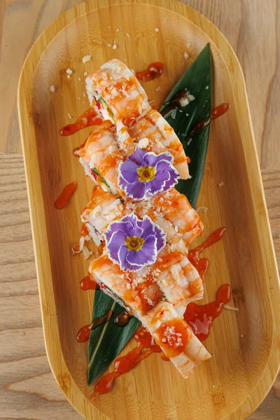 Karidesle doldurulmuş suşi ruloları ve ahşap bir tabakta sosla servis edilir. Yemek, yenilebilir çiçek ve yapraklarla süslenerek, yiyeceklerin sunumunu vurguluyor..