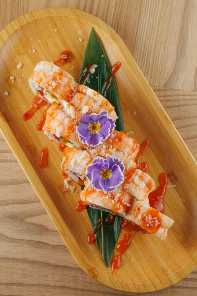 Suşi rulosu ahşap bir tabakta sosla süslenmiş ve yenilebilir çiçeklerle süslenmiştir. Bu yemek bir yemek ortamında lezzet ve sunum karışımı sergiler..