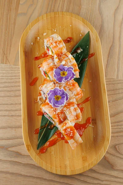 Suşi ruloları ahşap bir tabak üzerine serpiştirilir. Yenilebilir çiçekler yemeğe renk ve detay ekler. Bu yemek bir restoran ortamında Japon mutfağını ön plana çıkarıyor..