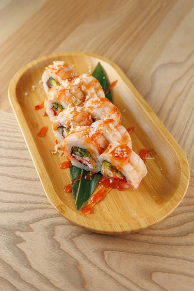 Ahşap bir tabakta çeşitli dolgularla renkli bir suşi rulosu sunulur. Sos üzerine serpilmiş, tabağın görünüşünü güçlendiriyor. Sahnede kırsal görünüm için ahşap bir masa var..