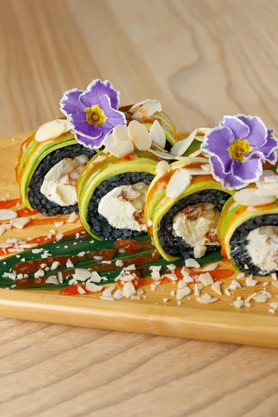 Suşi ruloları ahşap bir tabakta, tavuk dolması, siyah pirinç ve renkli süslemelerle düzenlenir. Tabakta yenilebilir çiçekler ve sos yağmurları yer alıyor..