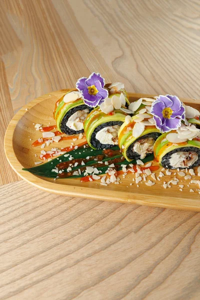 Suşi ruloları ahşap bir tabağa konur. Ekmeğin üzerinde yeşil katmanlar, bademler ve narin çiçekler var. Bir sos yağmuru tabağa renk ve detay ekler.