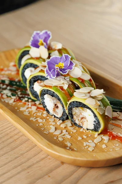 Suşi hamuru ahşap bir tabakta güzelce sıralanmış. Tabakta çiçek ve badem gibi renkli malzemeler ve süslemeler yer alıyor. Çok canlı bir sunum içeriyor..