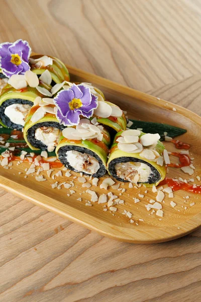 Deniz mahsulleriyle dolu suşi ruloları bambu tepsinin üzerinde oturur. Çiçeklerle ve dilimlenmiş bademlerle kaplı tabak renkli sunumlar ve ahşap bir yüzeyde taze malzemeler sergileniyor..