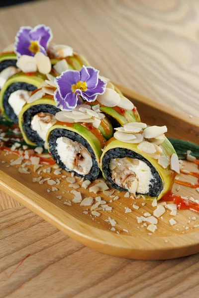 Krem peynirli, bademli ve yenilebilir çiçeklerle kaplı suşi ruloları ahşap bir tabakta oturur. Dekor, yiyeceklerin sunumunu geliştiren çağdaş bir yemek alanını gösteriyor..
