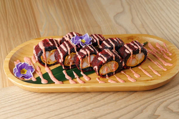 Taze suşi ruloları ahşap bir tabakta, sosla kaplı ve yenilebilir çiçeklerle süslenmiş. Bu yemek Japon mutfağında tipik bir tat ve renk karışımı sergiliyor..