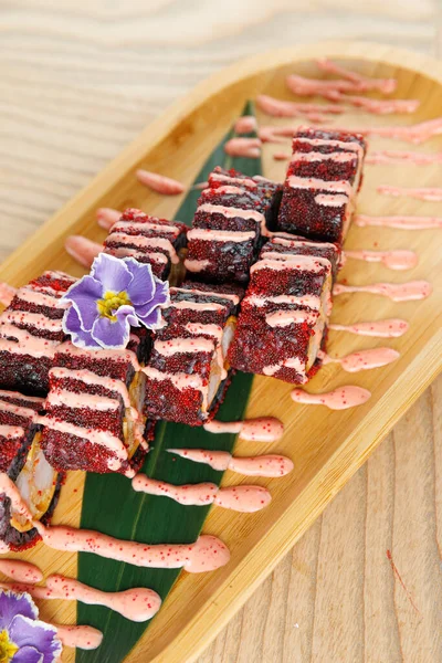 Kırmızı ve pembe katmanlı parlak suşi ruloları bambu tabakta düzenlenir. Bir çiçek dekoratif bir dokunuş eklerken, sos çiselemeleri görsel çekiciliği artırır..