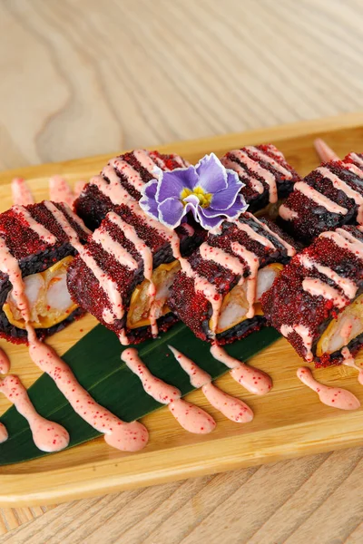 Suşi ruloları bambu tabakta güzelce düzenlenmiştir. Ekmekler canlı kırmızı malzemeler ve üzerine mor bir çiçek süslemesi serpiştirilmiş bir sos serpiştiriyor..
