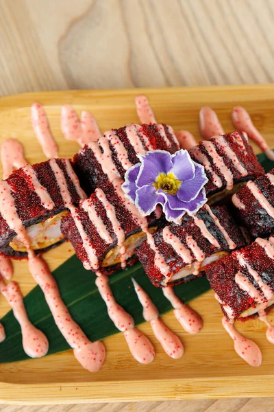 Suşi ruloları ahşap bir tepsiye güzelce yerleştirilir, pembe sos ve mor bir çiçekle süslenir. Tabakta Japon mutfağını vurgulayan çeşitli doku ve renkler yer alıyor..
