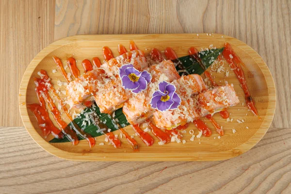 Tabak, ahşap bir tepside bitkilerle ve çiçeklerle kaplı somon balığı içeriyor. Sos yiyeceklerin etrafında çekici bir tasarım oluşturuyor. Bu tabak farklı mutfak geleneklerinden elementleri birleştiriyor..