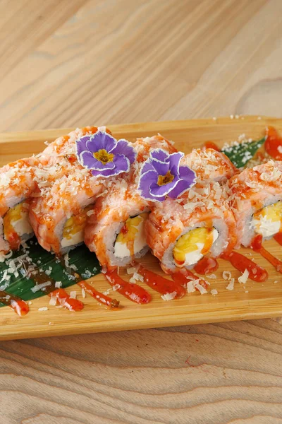 Parlak suşi ruloları ahşap bir tabakta düzenlenir. Tabakta renkli malzemeler, sos yağmurları ve dekoratif çiçekler yer alıyor. Lezzetli bir yemek için mükemmel..