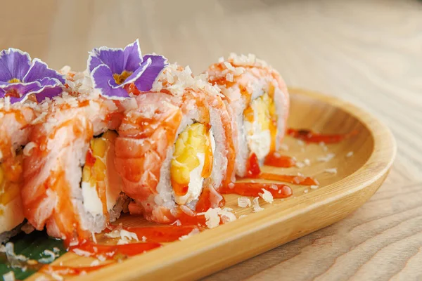 Pirinç, somon ve diğer malzemelerden yapılmış renkli suşi ruloları. Ahşap bir tabakta, sosla ve yenilebilir çiçeklerle düzenlenmişler. Burası bir yemek salonu..