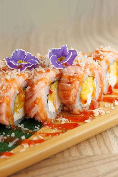 Suşi ruloları ahşap bir tabakta sunulur. Ekmekler somon ve yumurta içeriyor, üzerinde de yenilebilir çiçekler ve sos serpiştirilmiş. Renkli bir Japon mutfağı sergileniyor..