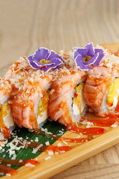 Somon ve mangoyla dolu suşi ruloları ahşap bir tabakta ustalıkla düzenlenir. Tabakta renkli süsler ve soslar yer alıyor, canlı bir sunum vurgulanıyor..