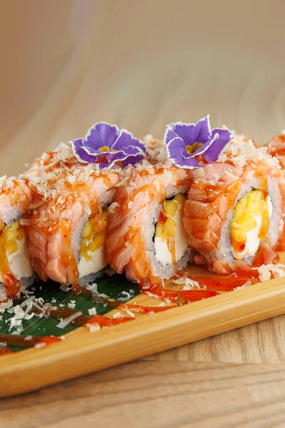 Somon ve krem peynirle dolu suşi ruloları ahşap bir tabakta oturur. Renkli çiçekler, yemek sunumunda lezzet ve yaratıcılığın bir karışımını sergileyerek tepeyi süslüyor..